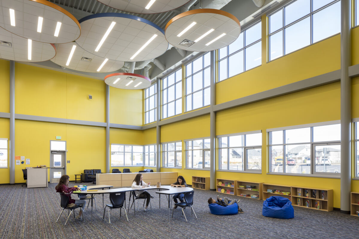 Learning space with yellow walls, large two‑story windows, circular acoustic light panels on the ceiling, bookshelves along the perimeter, and tables and seating areas on patterned carpet flooring.