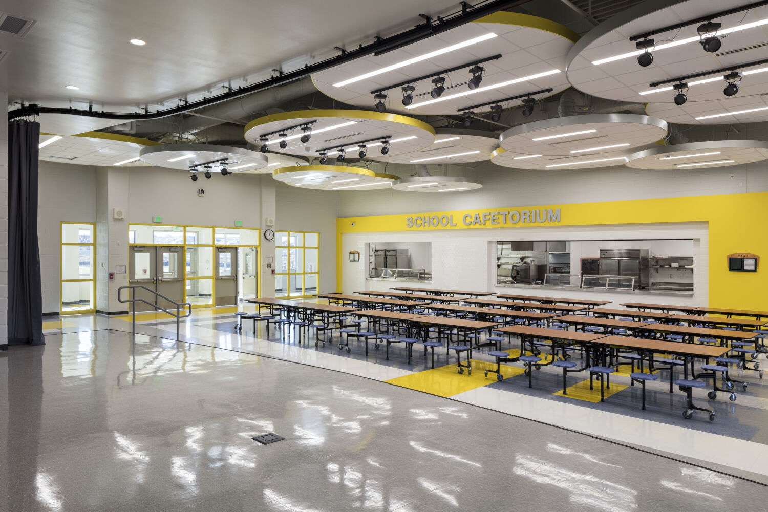 Cafetorium with folding cafeteria tables, yellow accent wall around the serving line, polished flooring, circular ceiling light panels, and multiple doors along the back wall.