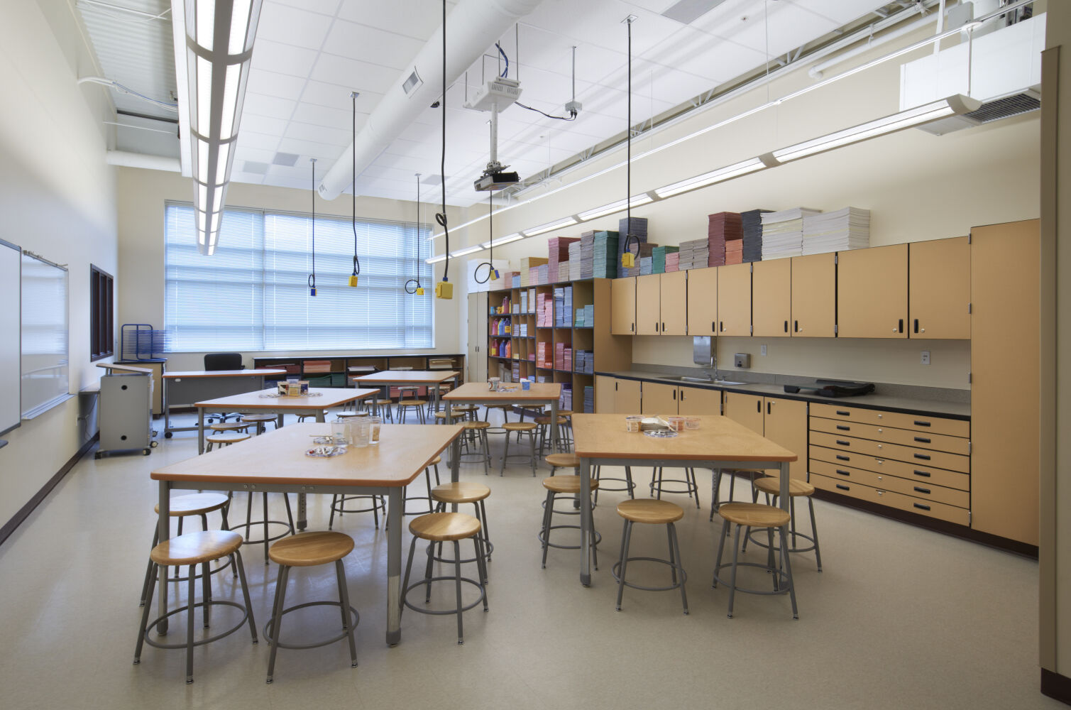 Art classroom with wood worktables and stools, storage cabinets, open shelving with colored paper stacks, large windows with blinds, tile flooring, and ceiling‑mounted utility reels.