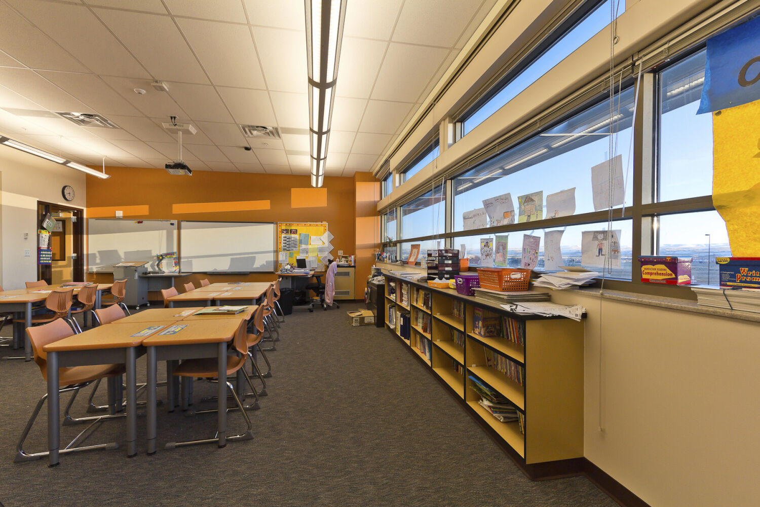 Classroom with grouped student desks and chairs, long horizontal windows, book shelving along the wall, orange accent wall near the whiteboard, and carpeted flooring.