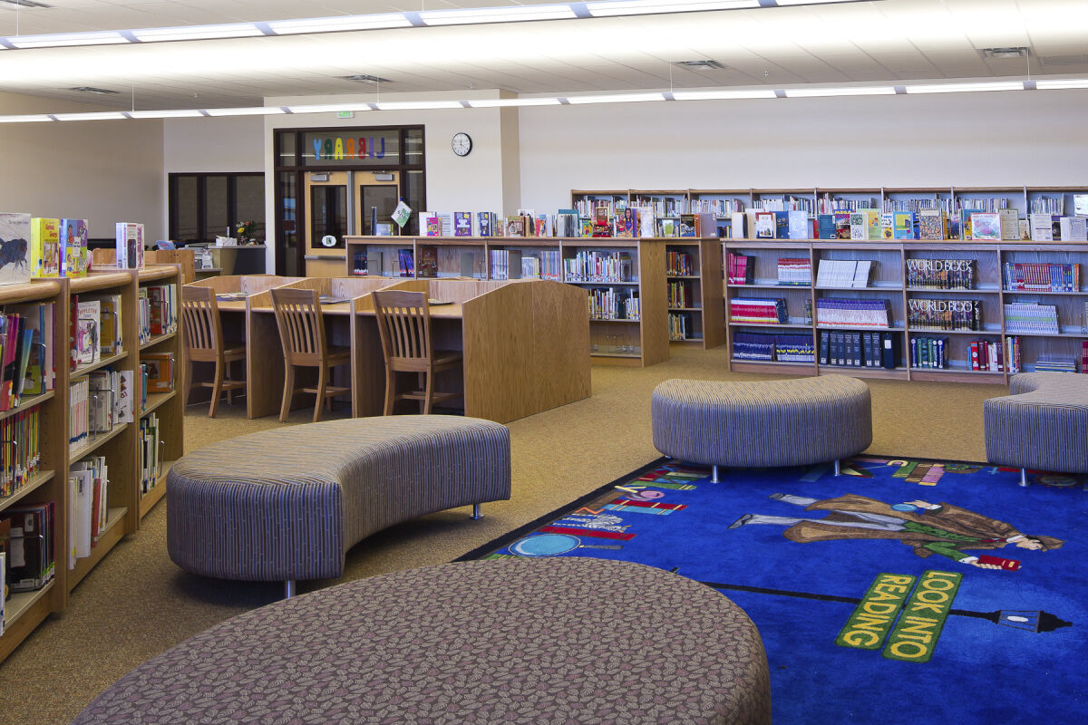 Library with curved lounge seating, wooden bookshelves, a blue story‑time rug, study tables with chairs, and bulletin boards along the back wall.