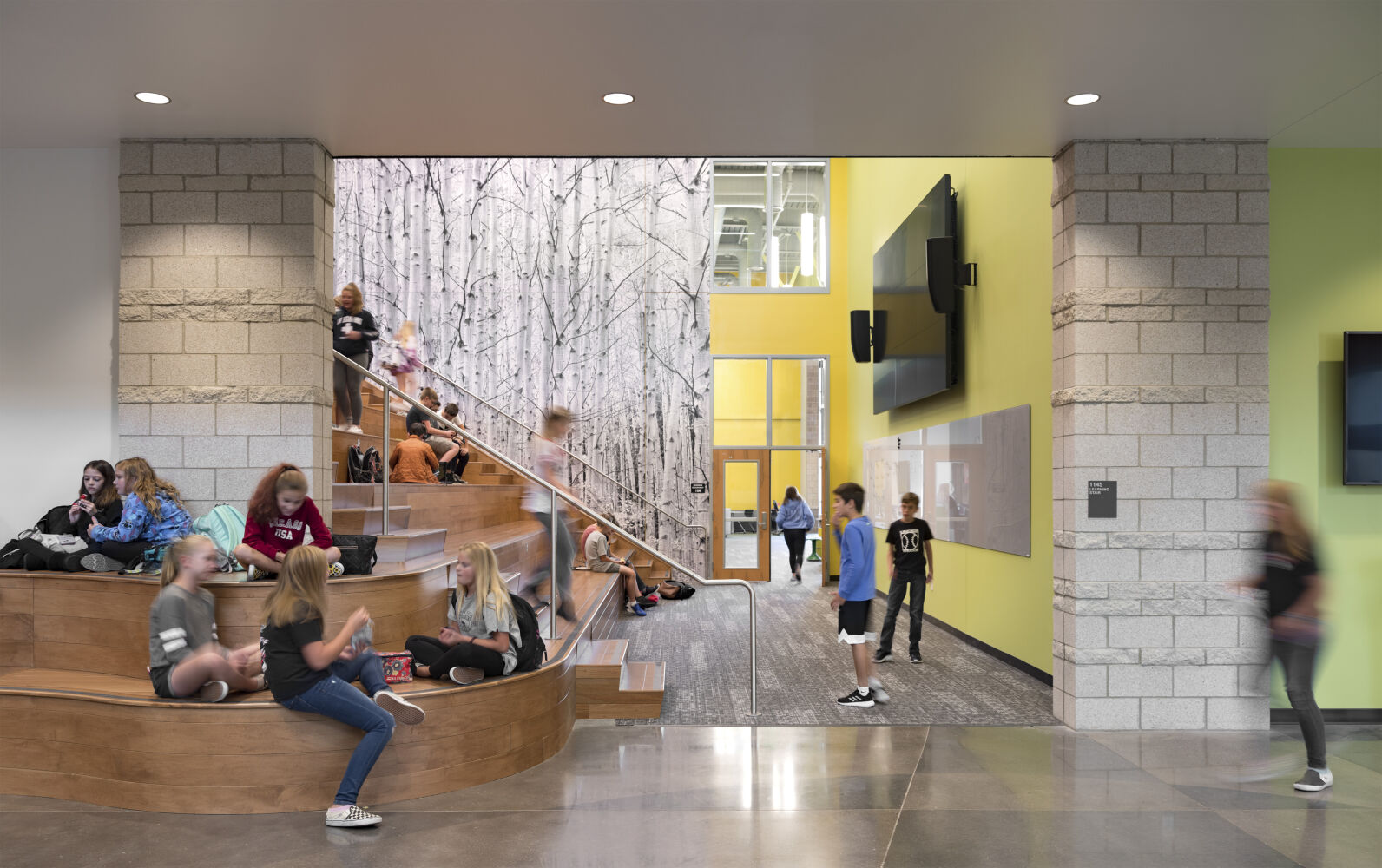 Learning stair with tiered wooden seating, adjacent birch‑forest wall mural, polished concrete flooring, yellow accent wall, and overhead lighting within a double‑height corridor.