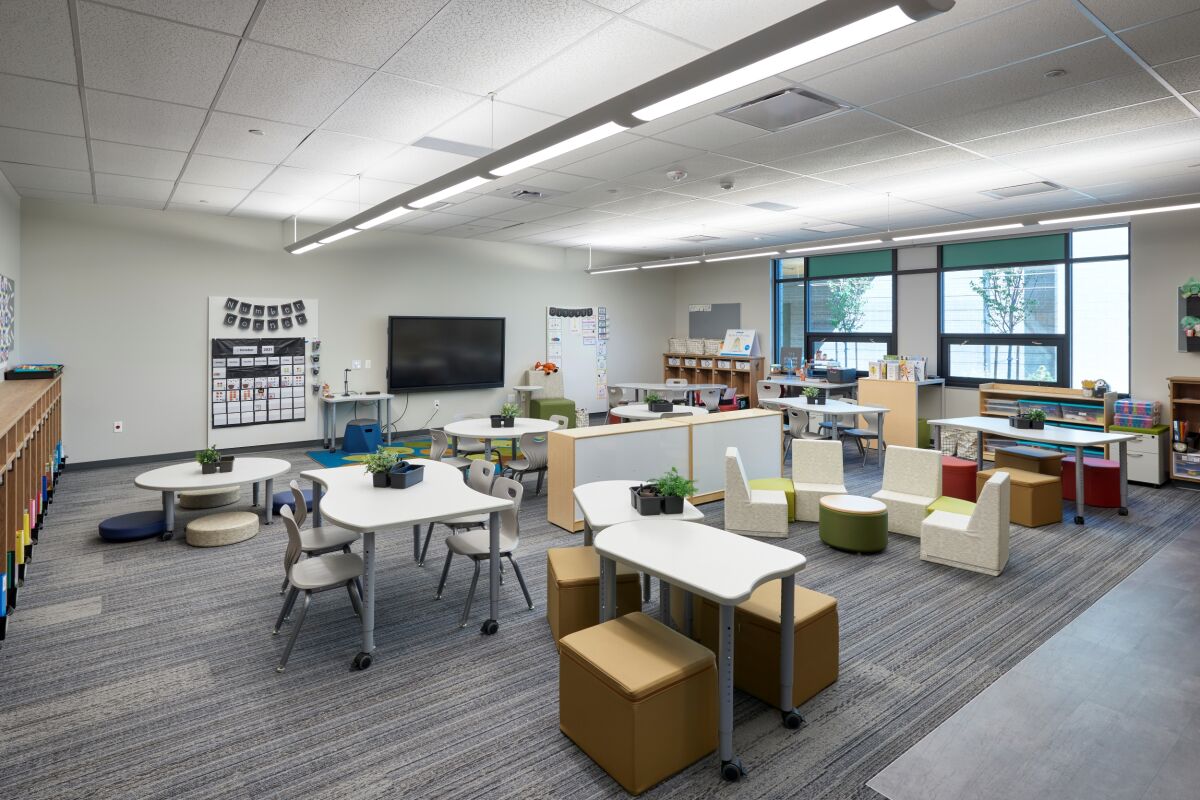 Classroom with flexible seating including movable tables, stools, and soft seating, carpet tile flooring, storage shelves with learning materials, large windows, and a wall‑mounted interactive display.