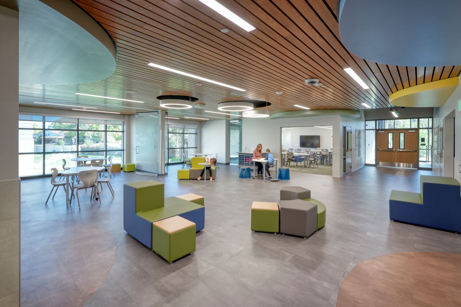 Collaboration area with movable soft seating in green and blue tones, round tables and stools, wood‑slat ceiling with circular light fixtures, and large windows overlooking an outdoor area.