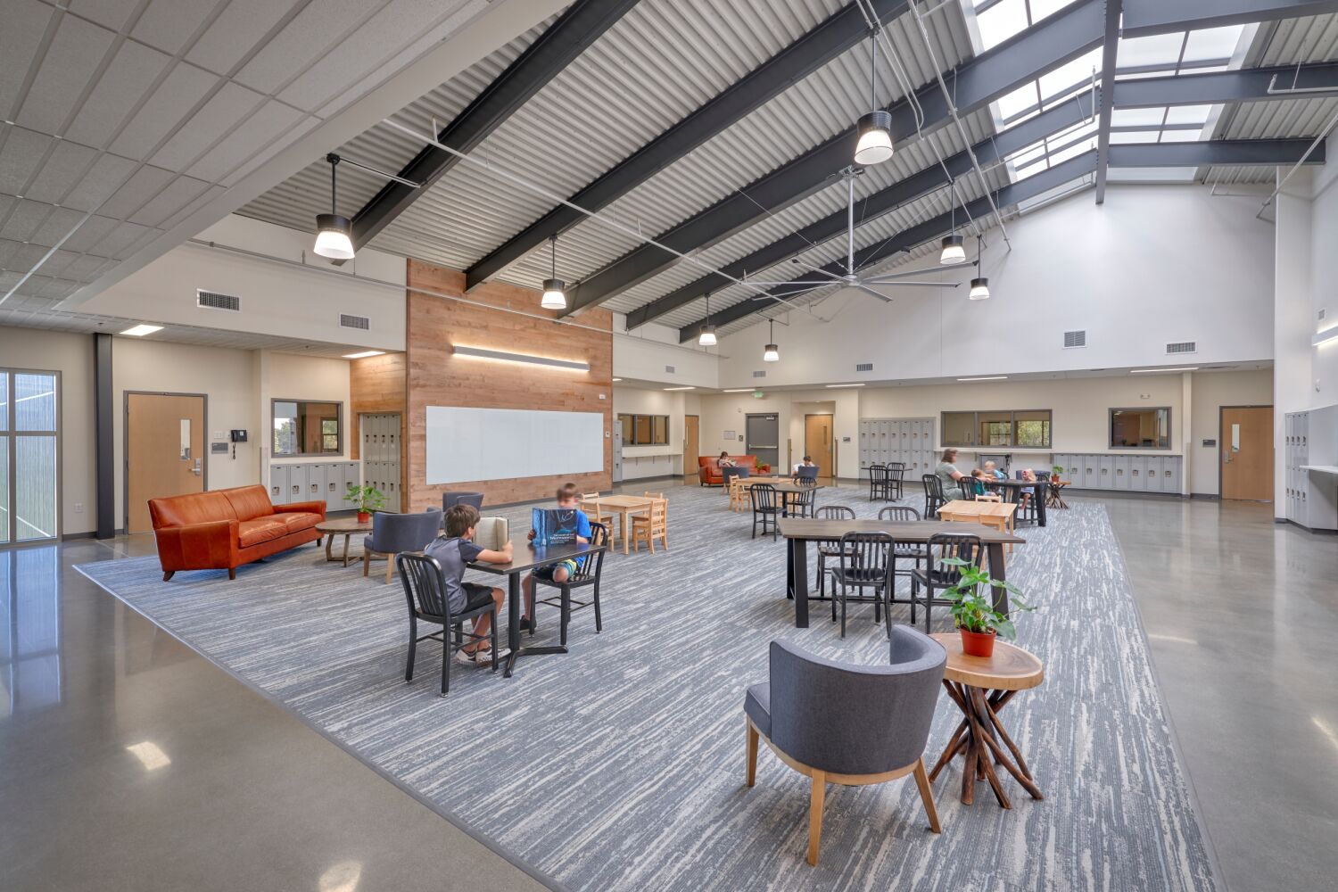 Great Work Montessori PK–5 School’s learning commons featuring skylights and exposed structural beams, lockers along the walls, a variety of tables and seating areas on an area rug, and large windows bringing in natural light.