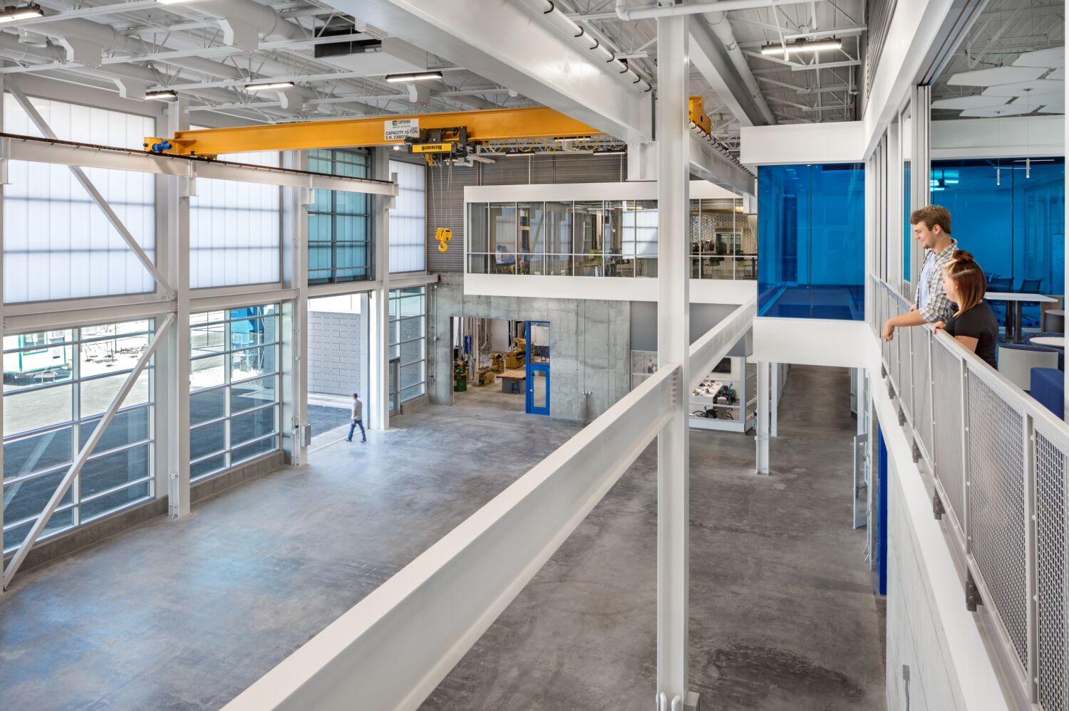 High-bay lab featuring tall translucent wall panels, exposed structural steel, an overhead yellow crane system, large glass garage doors, and an upper-level walkway with metal railing overlooking the open lab floor.