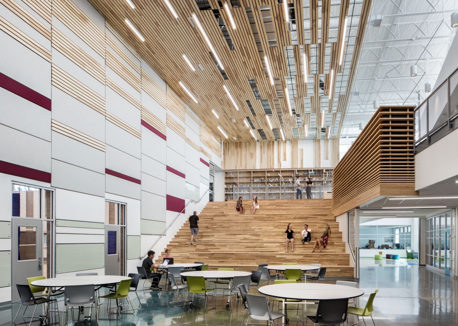 Pathways Innovation Center commons featuring wide tiered wood learning stairs, round tables and chairs in the foreground, a dramatic ceiling with suspended wood slats and linear lighting, and glass-walled rooms at the upper and lower levels.