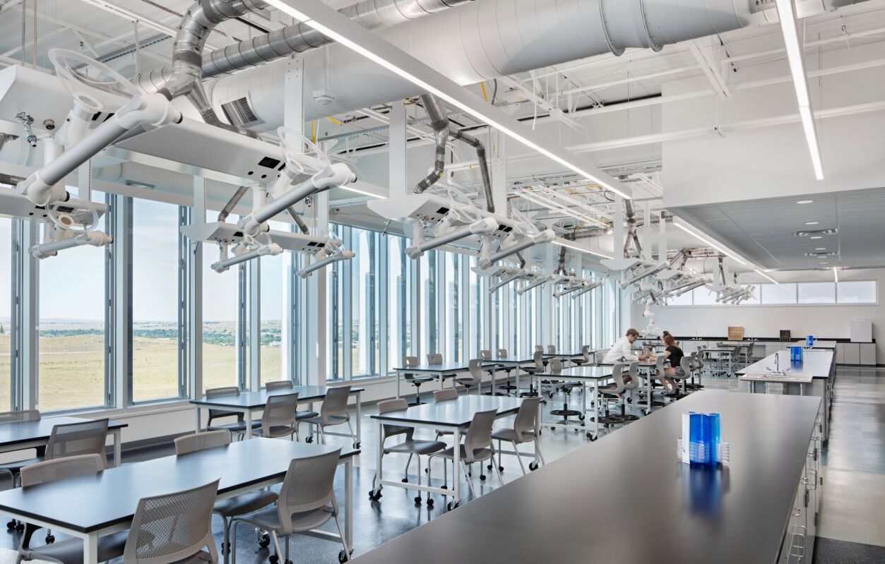 Pathways Innovation Center STEM lab featuring long worktables with stools, overhead ceiling‑mounted equipment arms, polished concrete floors, expansive floor‑to‑ceiling windows with views of open fields, and exposed ductwork and lighting.