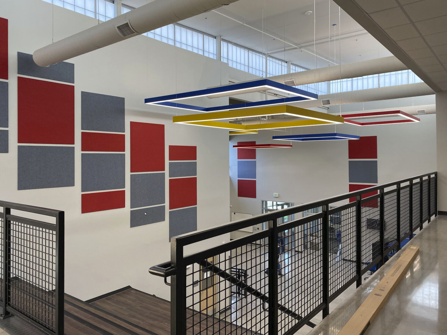 Upper-level overlook of the commons featuring red and gray acoustic wall panels, geometric ceiling lights, metal railing, and clerestory windows.