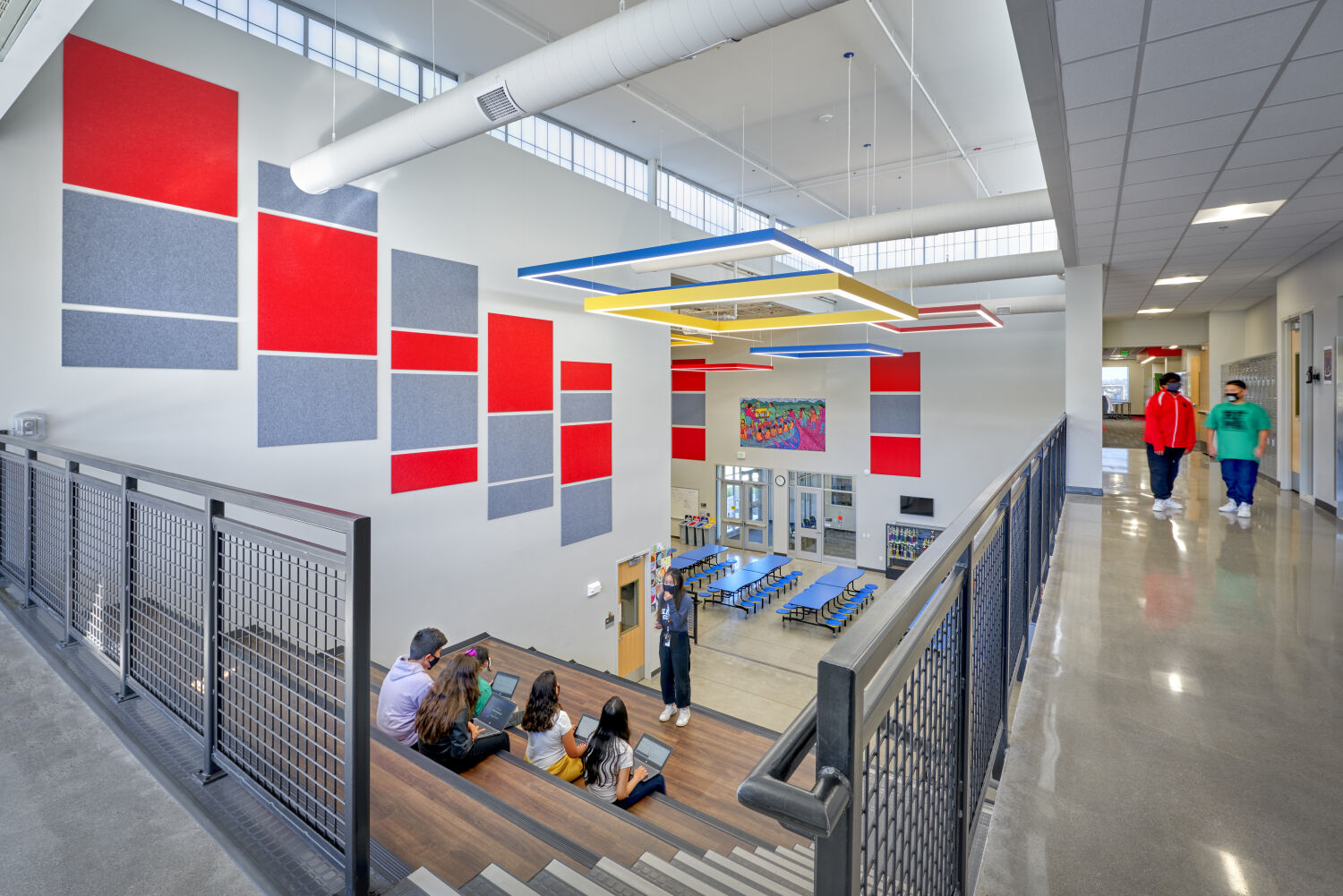 Learning stairs and cafeteria commons with colorful wall panels, geometric ceiling lights, metal railings, polished concrete floors, and high clerestory windows.