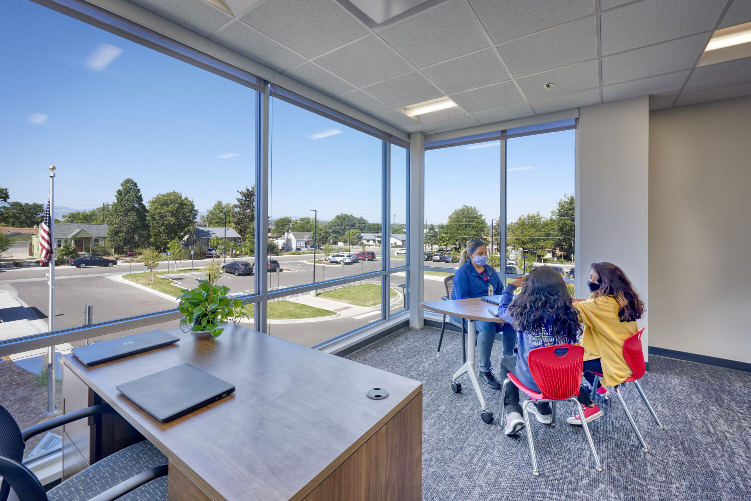 View of a conference room with a wood desk, red student chairs, carpet flooring, and large corner windows overlooking the surrounding neighborhood.