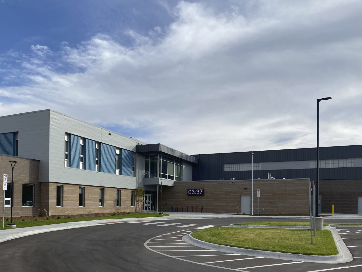Ricardo Flores Magón Academy main building with brick and blue-accent façades, a covered glass entry area, digital time display, surrounding parking loop with marked lanes, street lighting, and landscaped grass under a partly cloudy sky.
