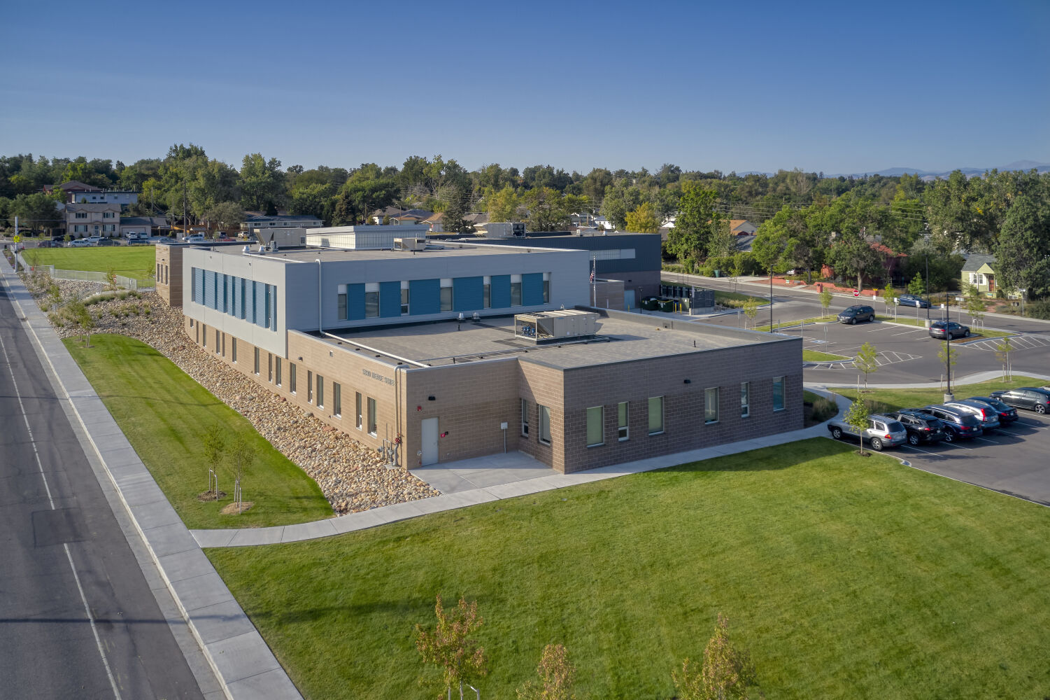 Ricardo Flores Magón Academy showing the classroom wing with brick and blue-accent façades, surrounding concrete walkways, a large green lawn, adjacent parking lot, and nearby residential neighborhood.