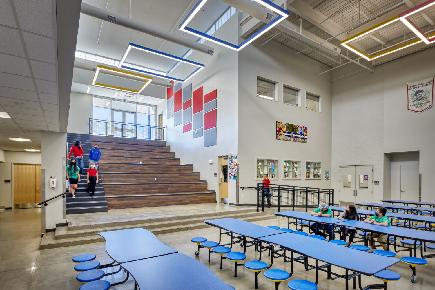 Ricardo Flores Magón Academy learning commons featuring wide tiered wood stairs, cafeteria tables with round stools, colorful wall panels, large ceiling light fixtures, polished concrete flooring, and an upper walkway with railing.