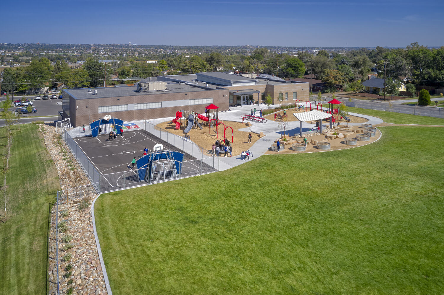 Ricardo Flores Magón Academy showing multiple playground areas with play structures, a paved play court, landscaped edges with rockwork, and the main school building set within a surrounding neighborhood.