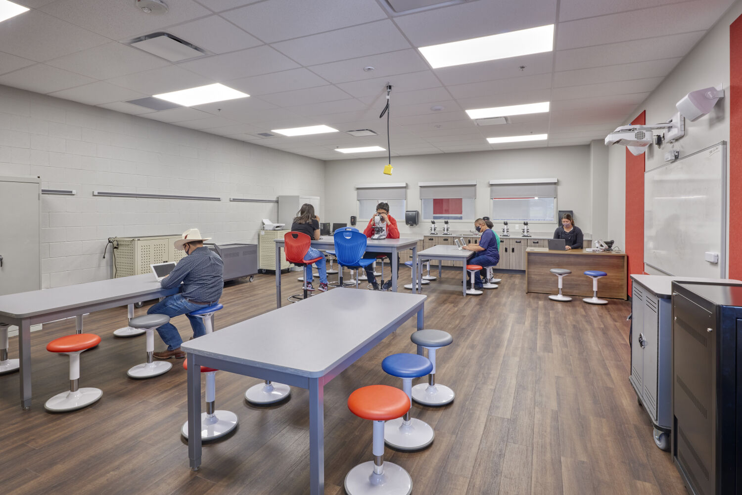 Science classroom with gray work tables, colorful round stools, wood flooring, whiteboard wall, storage cabinets, and microscopes arranged along the back counter.