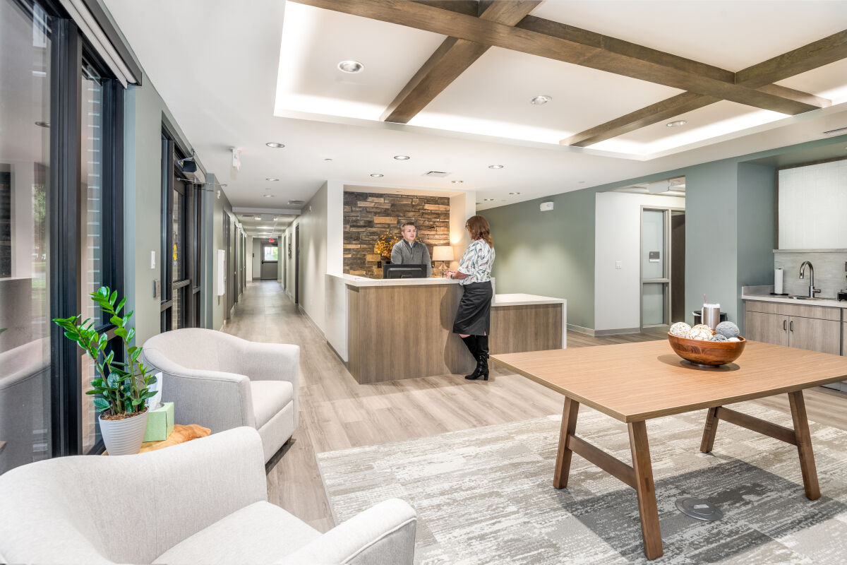 Reception lounge with exposed wood ceiling beams, a stone feature wall, wood flooring, neutral upholstered seating, large windows, and a central wood table.