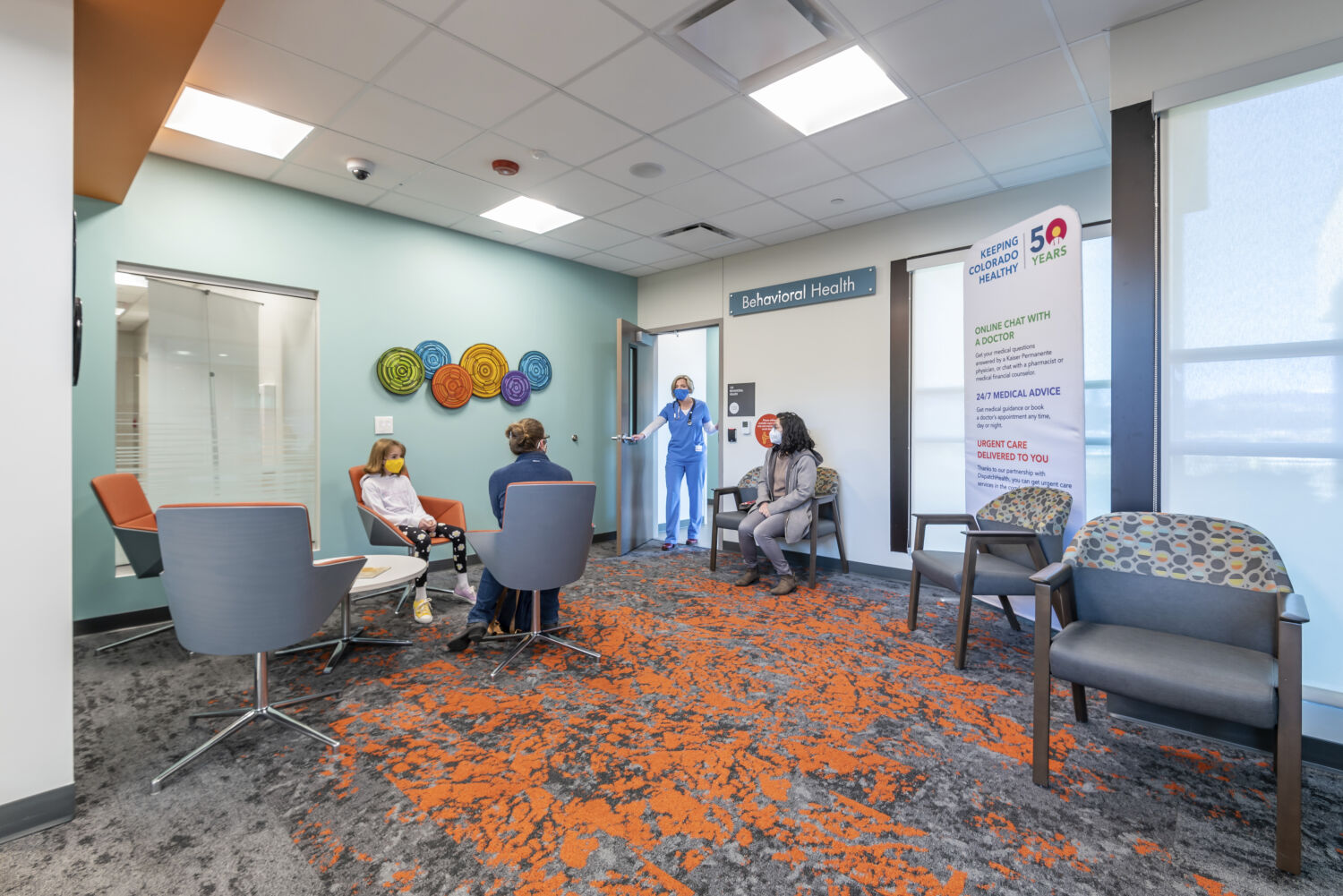 Behavioral health waiting area with colorful circular wall art, gray and orange seating, patterned carpet, teal accent wall, and a doorway marked “Behavioral Health.”