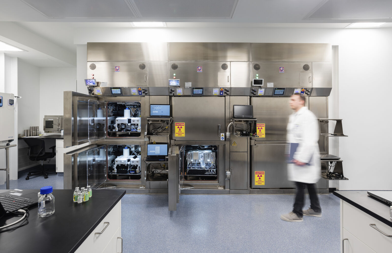 Hot‑cell suite with stainless steel shielded isolators, integrated glove ports, radiation‑warning labels, mounted touchscreens, lead‑glass viewing panels, and blue epoxy flooring.
