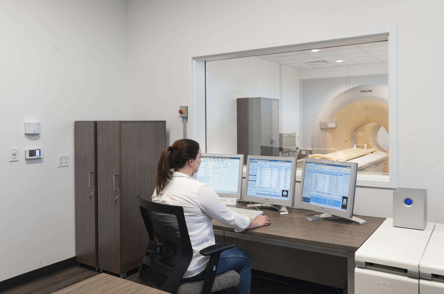 MRI control room with three monitoring stations, wood-finish cabinetry, a large interior observation window, and the MRI scanner visible in the adjacent suite.
