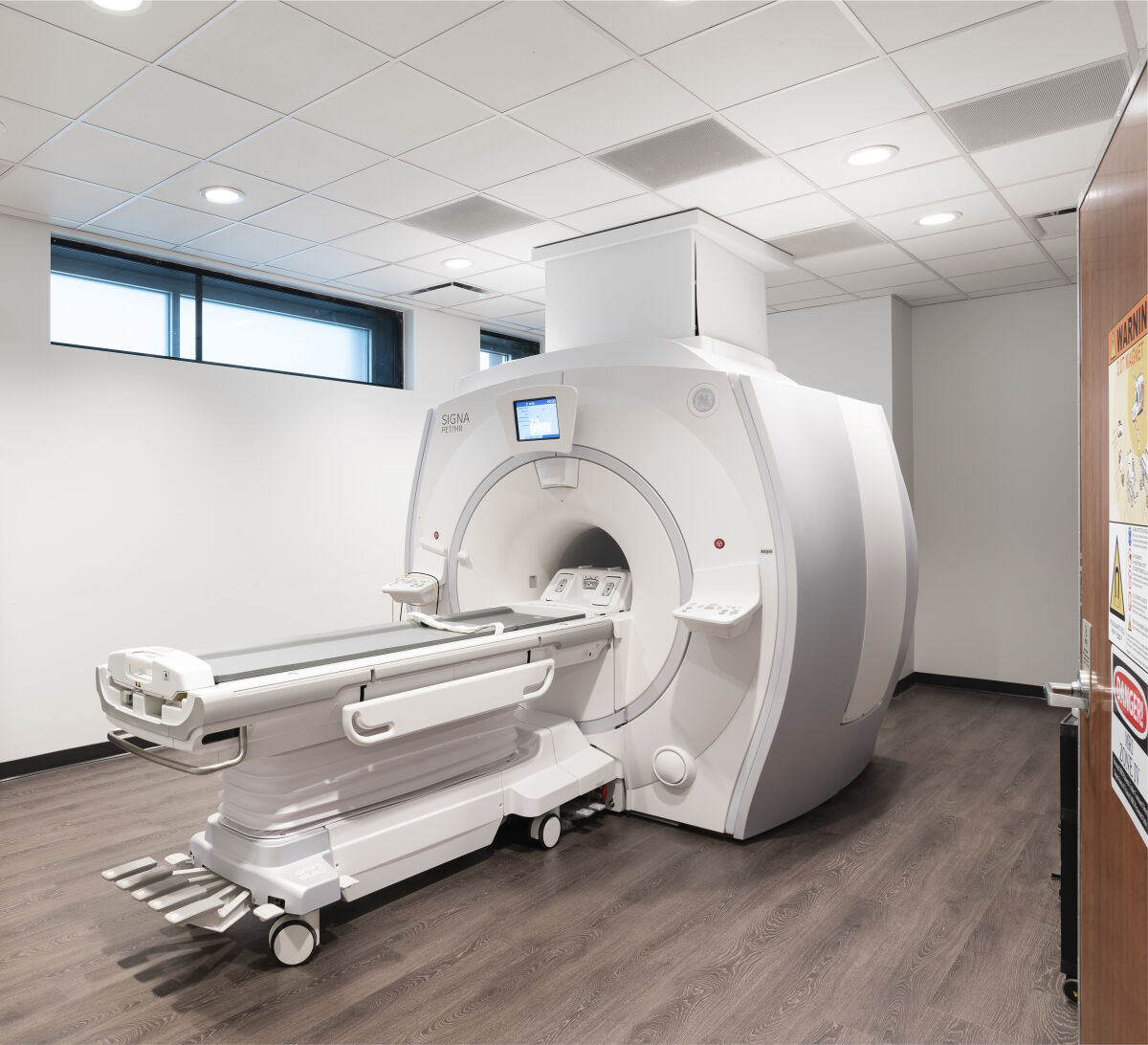MRI suite with a large 3T scanner, patient table on rails, wood-look flooring, acoustic ceiling panels, recessed lighting, and high windows along the wall.
