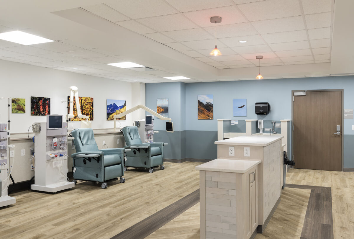 Dialysis area with teal treatment recliners, wall‑mounted equipment, landscape artwork, wood-look flooring, pendant lighting, and a central nurse station with white paneling.