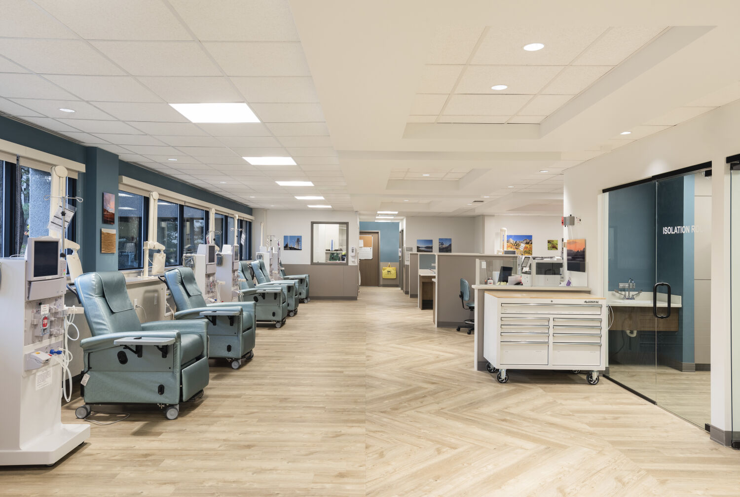 Dialysis treatment floor with teal recliner stations, wall-mounted equipment, wide windows, wood-look flooring, central workstations, and a glass-walled isolation room.