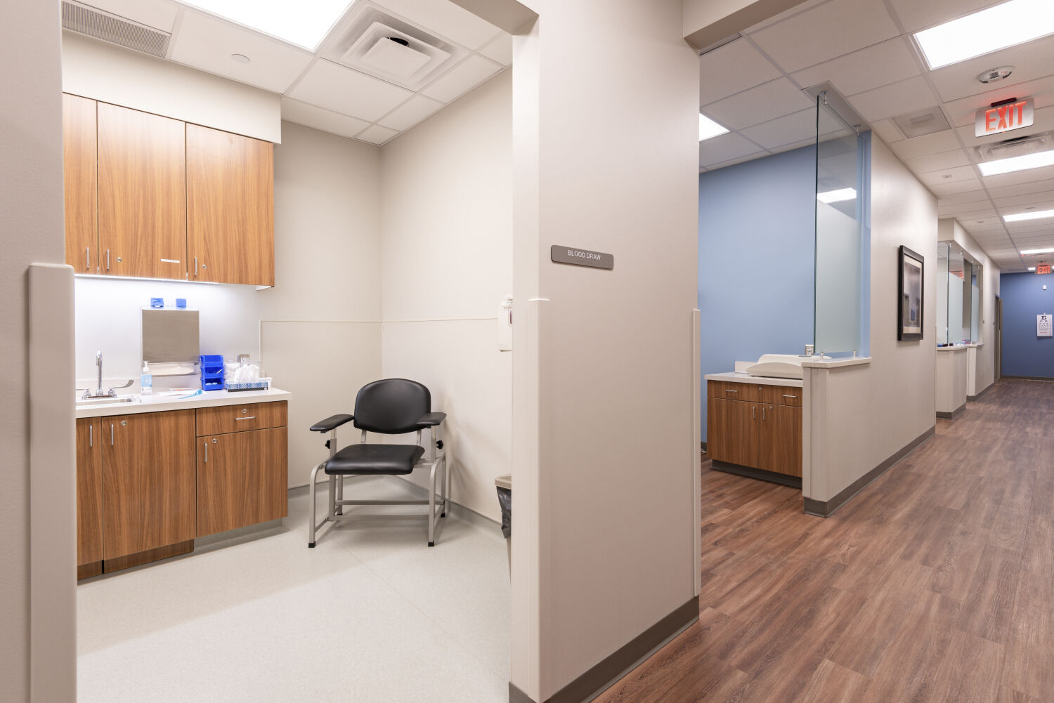 Clinical alcove with wood cabinets, a sink, and a black exam chair opening to a corridor with blue accent walls, wood-look flooring, and glass partitions at the nurse station.