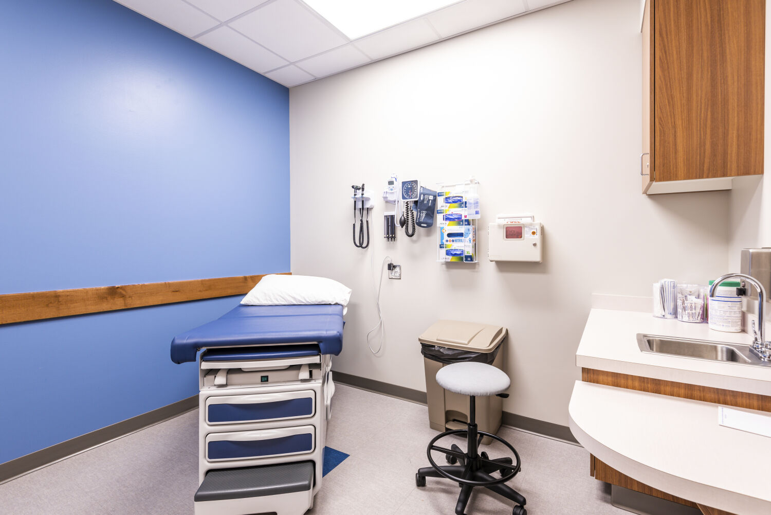 Exam room with a blue accent wall, medical exam table, wall‑mounted diagnostic equipment, wood cabinetry, a countertop sink, and neutral flooring.