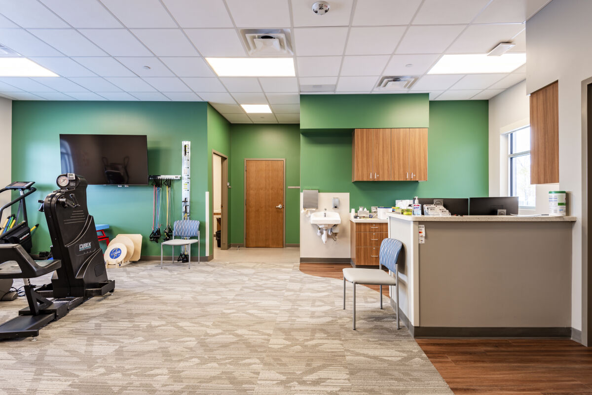 Rehab gym with exercise machines, wall‑mounted TV, green accent walls, wood cabinetry, a check‑in counter, and patterned carpet flooring.