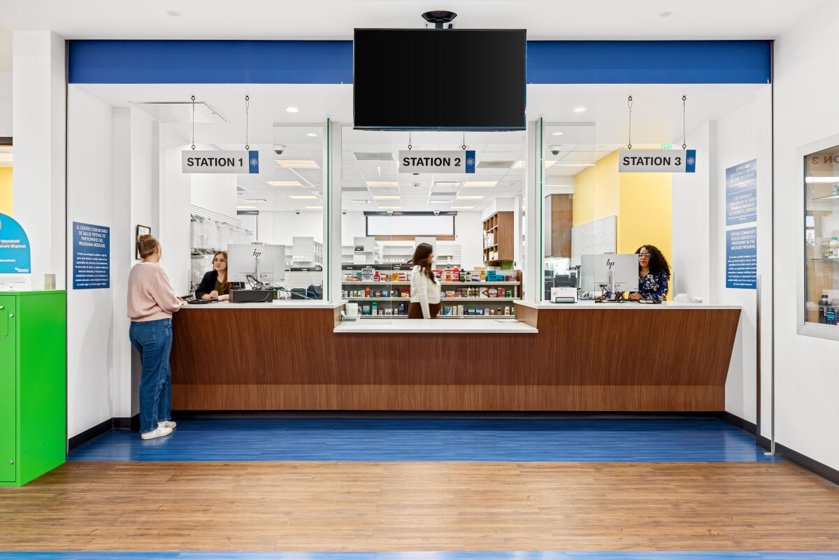 Pharmacy service counter with three labeled stations, wood cabinetry, overhead signage, and shelving stocked with medical and retail items behind a glass divider.