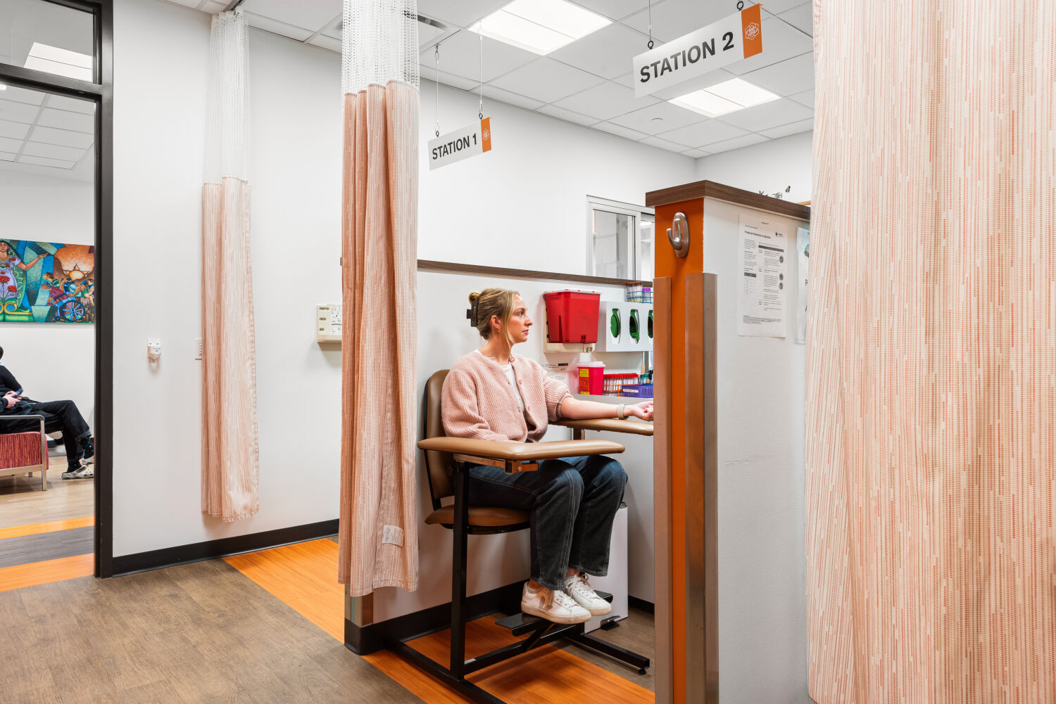 Phlebotomy draw station with a labeled overhead sign, privacy curtains, wood cabinetry, medical supplies, and a chair positioned beside a countertop pass-through window.