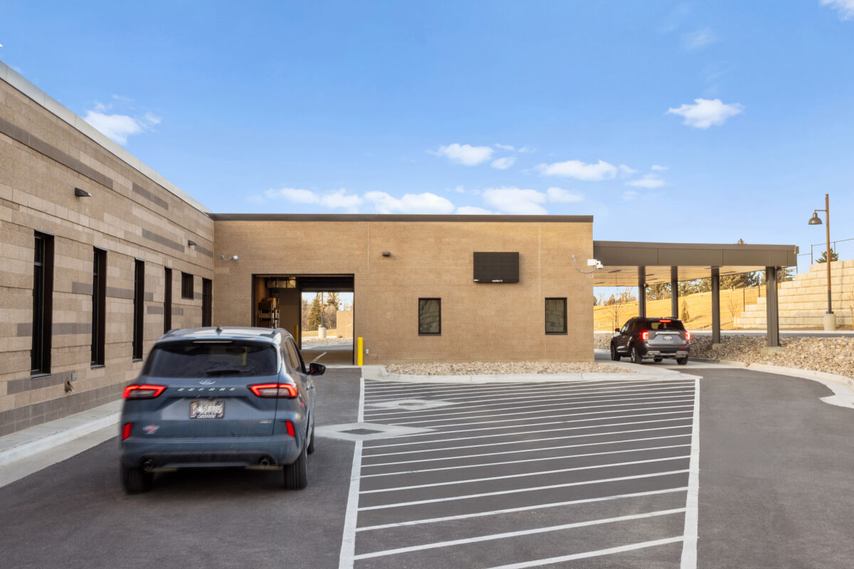 Drive‑through clinic area at the Casper–Natrona County Health Department with a covered canopy, brick service buildings, striped vehicle lanes, and adjacent parking access.