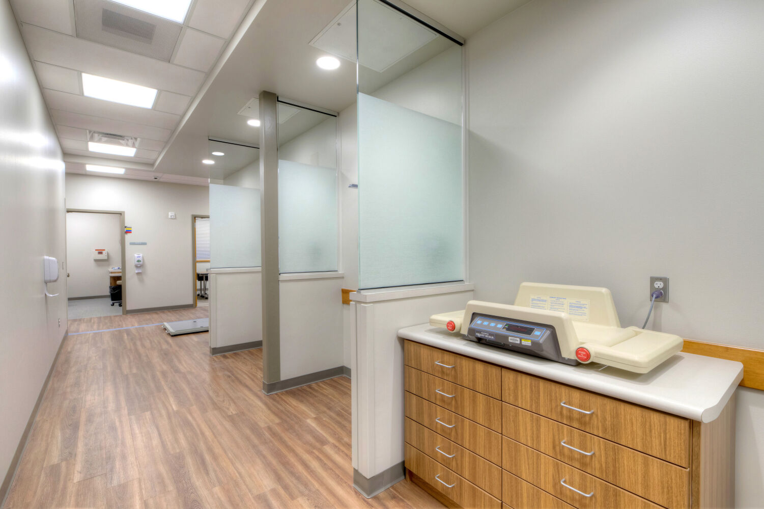 Clinical corridor with semi-private weighing alcoves featuring frosted glass panels, wood-look flooring, overhead lighting, and a countertop area with wood cabinetry and medical equipment.