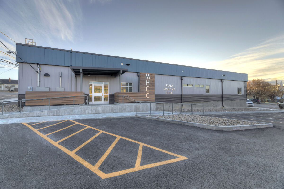 Exterior of the MHCC Rawlins Family Practice Clinic featuring metal siding, wood accent panels, a covered entry, accessible ramp, and striped parking area at sunrise.
