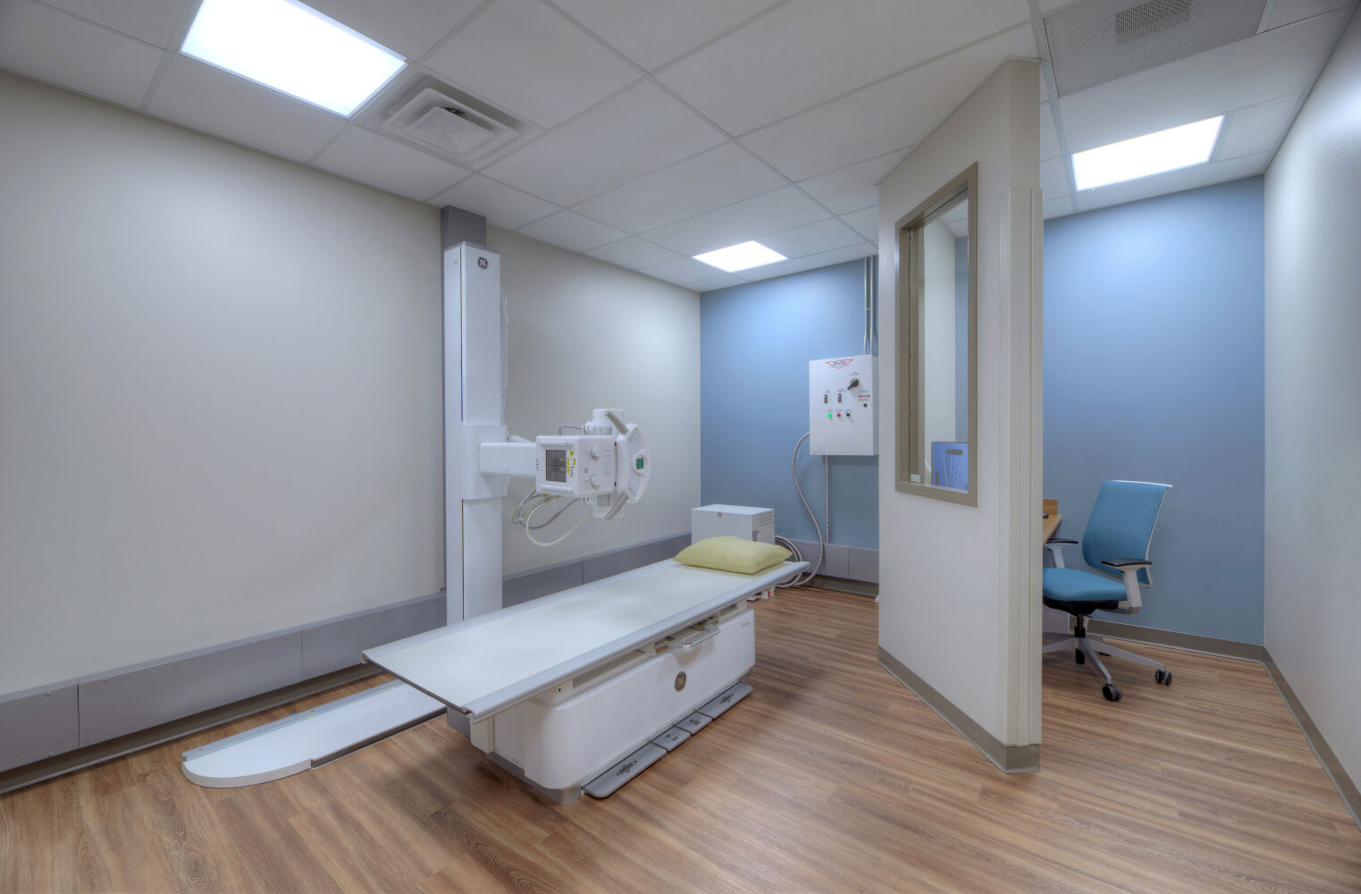 Imaging room with an exam table, wall-mounted radiology equipment, wood-look flooring, light-colored walls, and a teal accent wall beside a workstation with a rolling chair.