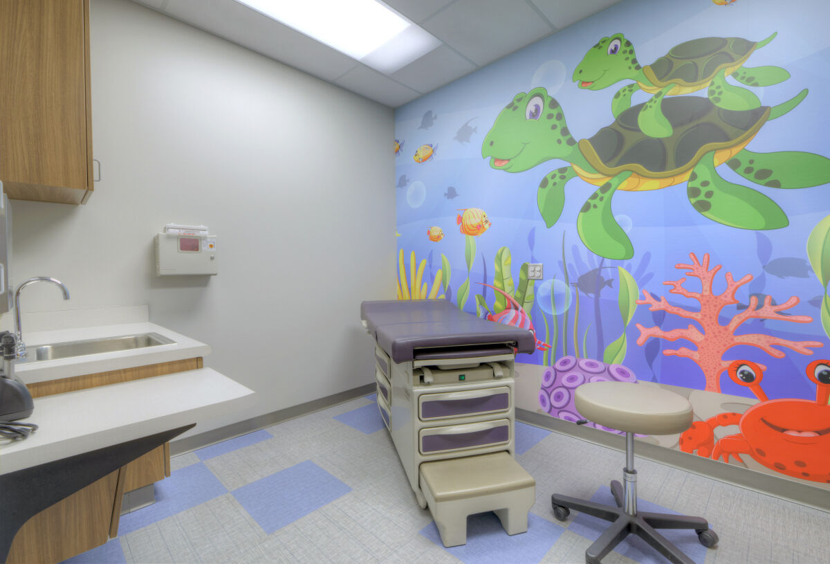 Pediatric exam room with a colorful underwater-themed mural, exam table, wood cabinetry with a sink, vinyl flooring in blue and gray tones, and overhead fluorescent lighting.