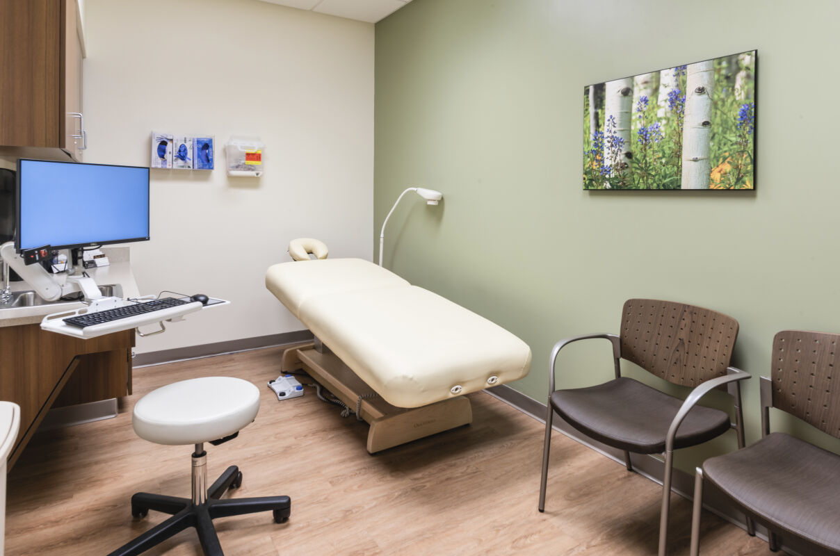 Exam room with a padded treatment table, wall-mounted computer workstation, wood cabinetry with a sink, green accent wall with nature artwork, and vinyl wood-look flooring.