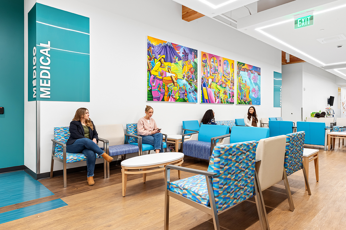Waiting area with turquoise seating, patterned chairs, wood flooring, and a series of colorful murals mounted on a white wall beneath linear ceiling lighting, with a teal “MEDICAL” sign to the left.