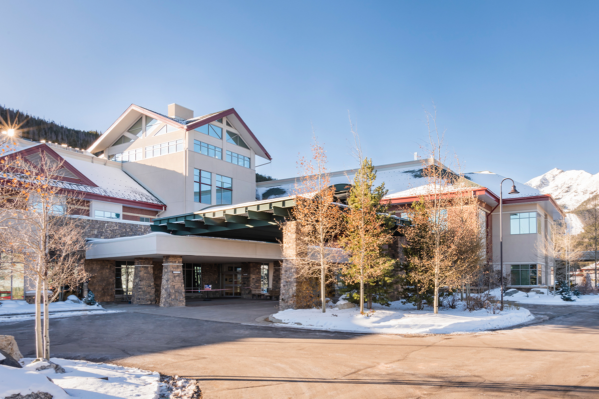 Exterior of the St. Anthony Summit Medical Center Emergency Department featuring a covered drop-off canopy, stone and stucco façade, large windows, and surrounding snow-covered landscaping with mountain views in the background.