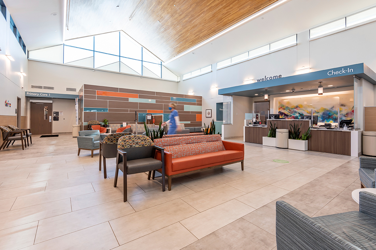 Lobby with colorful lounge seating, a blue check-in canopy, tiled flooring, a wood-paneled ceiling, clerestory windows, and a large geometric accent wall near the waiting area.
