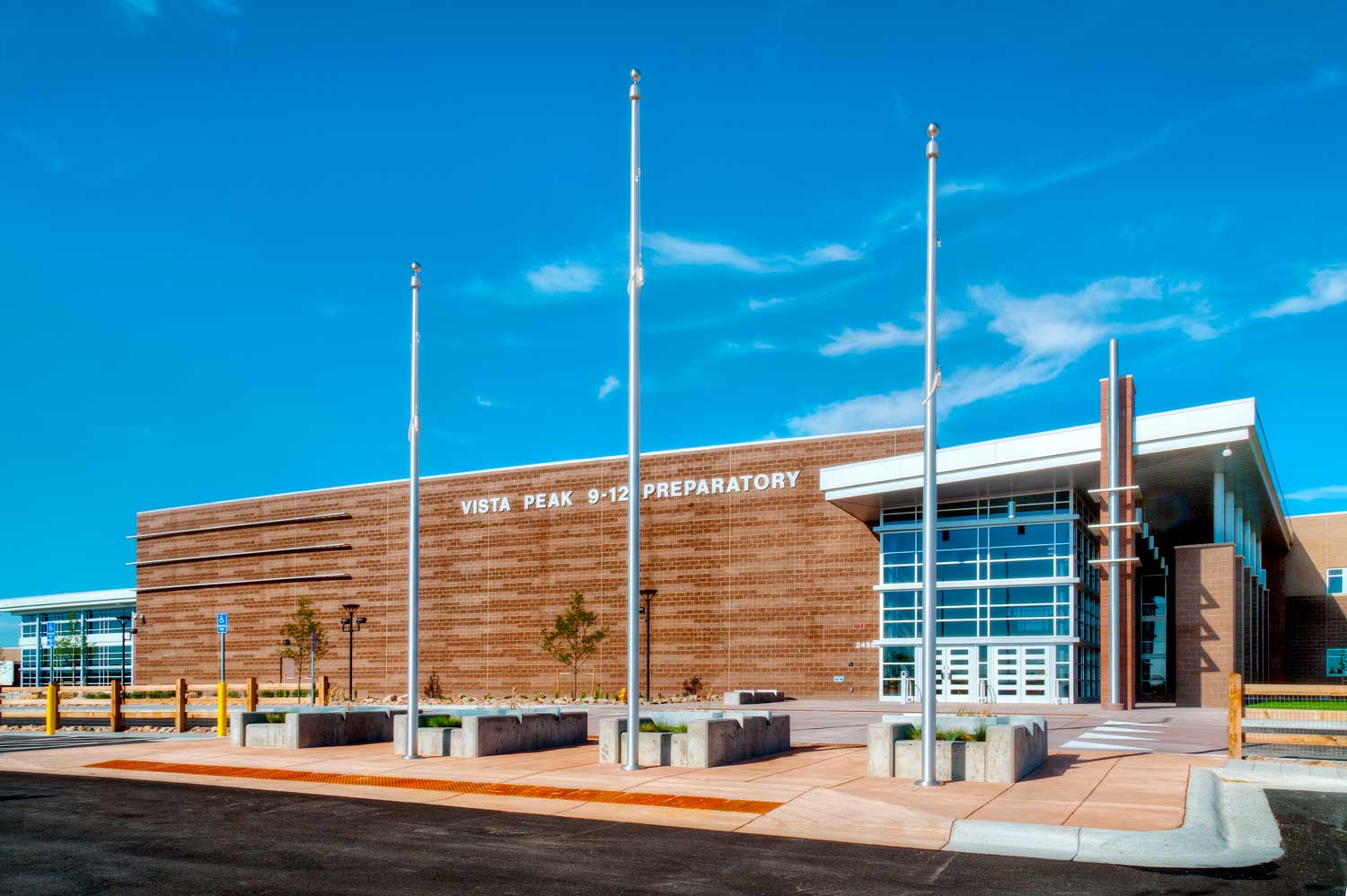 Exterior view of Vista Peak High School, showcasing a modern architectural design with a combination of brick and metal materials. The building features large windows and a grand entrance, set against a clear blue sky and surrounded by well-maintained landscaping.