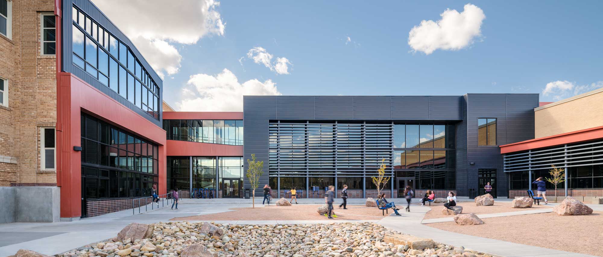 Exterior view of CCMS courtyard, featuring a modern school building with large windows, brick and metal accents, and an open outdoor gathering space. The courtyard is designed for student interaction and relaxation, with seating areas and pathways enhancing accessibility.