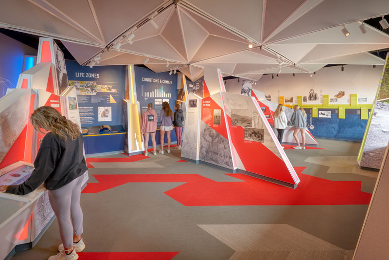Interior view of the Pikes Peak Visitor Center exhibit, featuring interactive displays and informational panels. The space highlights the natural and cultural history of the area, with modern lighting and a clean design that enhances the visitor experience.