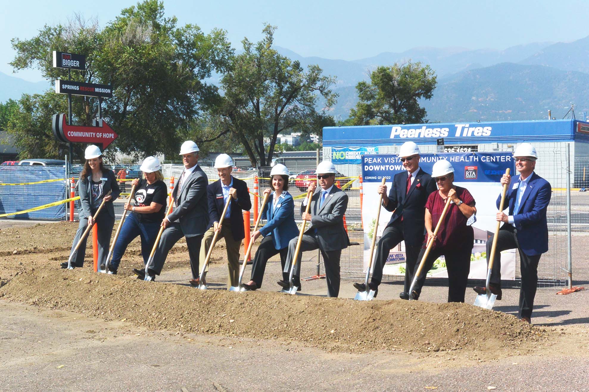 A group of people wearing hard hats prepare to shovel a pile of dirt as part of the groundbreaking ceremony